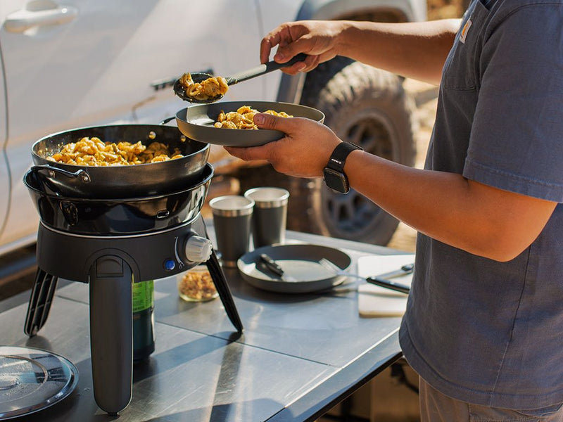 Load image into Gallery viewer, Person cooking on a portable Front Runner Safari Chef 30 HP gas barbeque, perfect for camping and outdoor cooking.
