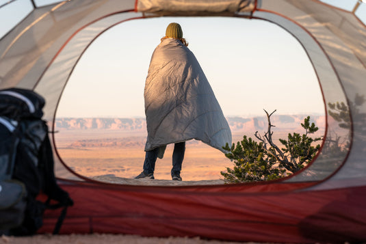 Person wrapped in Klymit Horizon Overland Blanket standing outside a tent with scenic desert landscape in the background.