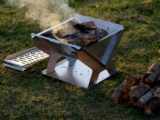 Portable stainless steel Front Runner BBQ/Fire Pit set up outdoors with smoldering wood and a grill plate on the side.