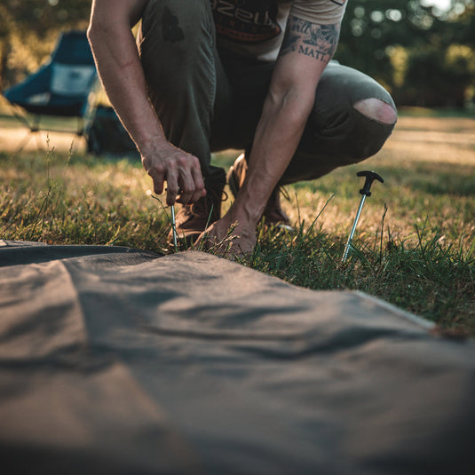 Person securing a Gazelle T4 Tent Footprint to the ground with a metal stake.