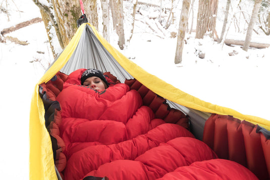 Person resting in a yellow and grey hammock outdoors with a red Klymit Insulated Hammock V Sleeping Pad, surrounded by snowy trees.