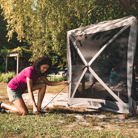 Alt text: "Woman setting up a Territory Tents 6-Sided Screen Tent outdoors on grass with trees in the background."