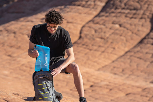 Man filling Klymit Echo 12L Hydration Pack with water while sitting on rocky terrain outdoors.