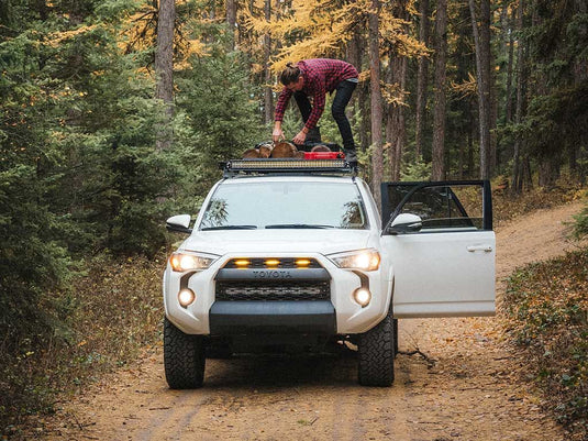 Person loading camping gear onto Front Runner Slimline II Roof Rack on a white Toyota 4Runner parked in a forest.