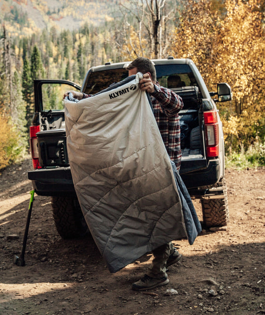Man unfolding Klymit Horizon Overland Blanket next to SUV in autumn forest setting