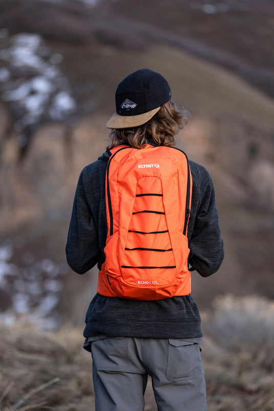 Person wearing Klymit Echo 12L Hydration Pack in orange with reflective stripes, outdoor setting.