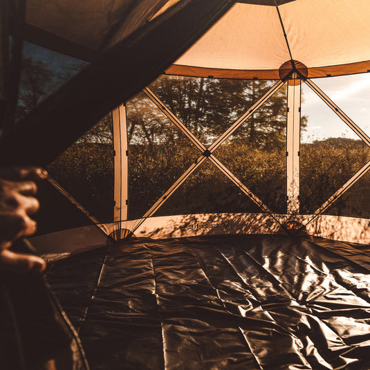 Alt text: "Interior view of a Gazelle Tents G6 6-Sided Gazebo showcasing the footprint flooring and screened panels with a forest background at sunset."