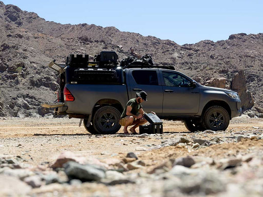 Alt text: "Person organizing gear beside a truck equipped with Front Runner Wolf Pack Pro Hi-Lid storage containers in a rugged outdoor setting."