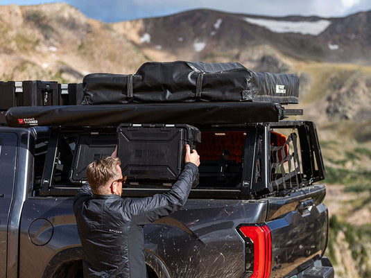 "Man installing Front Runner Pro Bed Rack Kit on Toyota Tacoma Double Cab with storage boxes and gear against mountain backdrop"