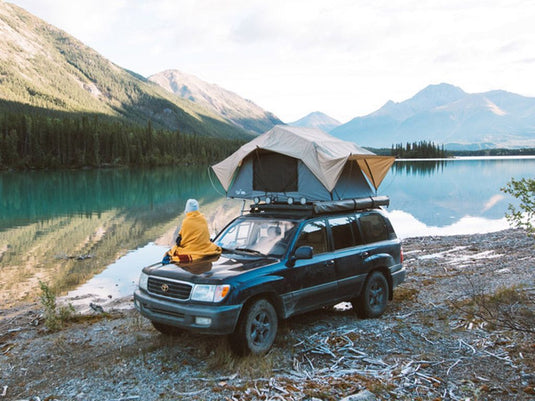 Person sitting on a Toyota 100 Series Land Cruiser with a Front Runner Slimline II Roof Rack and roof-top tent by a serene lake with mountains in the background.