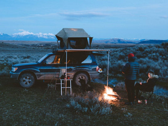 Toyota 100 Series Land Cruiser with Front Runner Slimline II Roof Rack and rooftop tent set up at a dusk campsite with two individuals by a campfire and mountain landscape in the background.