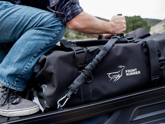 Man securing a black Front Runner Stratchit to a duffle bag on a vehicle roof rack.