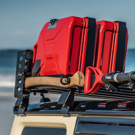 Front Runner Axe Bracket mounted on vehicle roof rack beside red fuel cans with ocean in the background