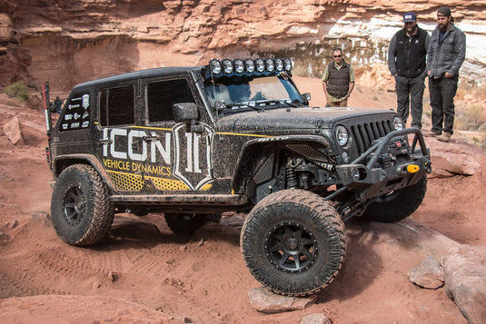 Jeep equipped with ICON Vehicle Dynamics Rebound wheels in Satin Black finish, showcasing off-road capability in a rocky desert terrain with people observing in the background.