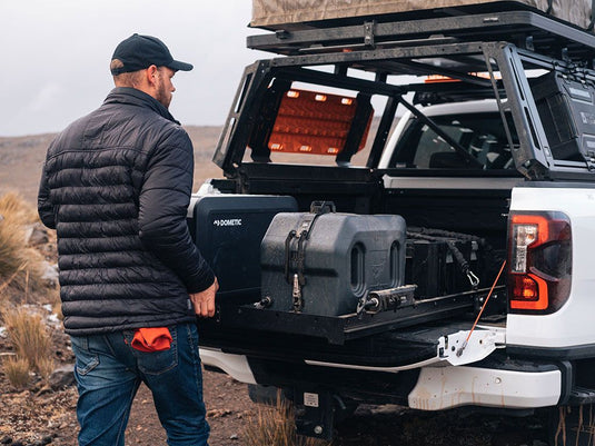 Alt text: "Man standing next to a Toyota Tacoma Double Cab with a Front Runner Pro Bed Rack Kit installed, featuring storage boxes and camping gear mounted on the rack in an outdoor setting."