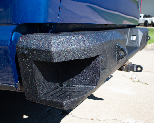 Alt text: "Close-up view of a textured Fishbone Offroad Pelican Rear Bumper installed on a 2015-Current Ford F-150, featuring cutouts and tow hook mounting points."