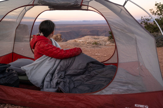 Person sitting in a tent at sunrise draped in a Klymit Horizon Overland Blanket, overlooking a scenic canyon.
