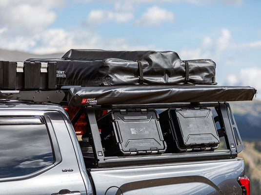 Alt text: "Front Runner Pro Bed Rack Kit installed on a Toyota Hilux Revo Double Cab 2016, equipped with gear storage boxes and roof-mounted cargo bag against a mountainous backdrop."