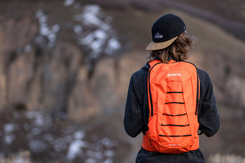 Load image into Gallery viewer, Person wearing Klymit Echo 12L Hydration Pack in bright orange color, designed for outdoor activities, with reflective stripes and branding visible.
