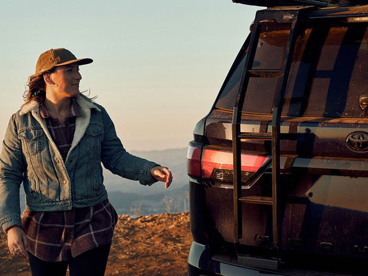 Woman standing next to a Toyota Sequoia with a Front Runner ladder attached to the rear, outdoors at dusk.
