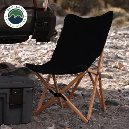 Alt text: "Overland Vehicle Systems Kick It Camp Chair with wooden legs and black fabric set up outdoors near a storage container and an off-road vehicle."