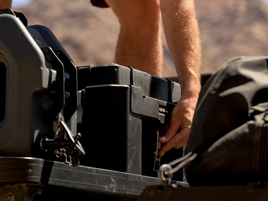 Alt text: "Close-up of a person attaching the Front Runner Wolf Pack Pro Rack Mounting Bracket MKII to a storage box mounted on a vehicle."